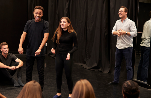 Two students perform a drama scene in front of a class. Their teacher directs from the side-lines.