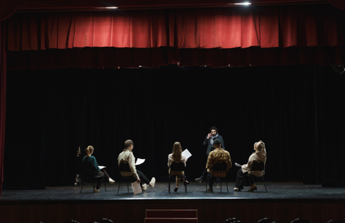 Five students are sat on a theatre stage holding scripts. Their teacher faces them, seemingly giving direction.